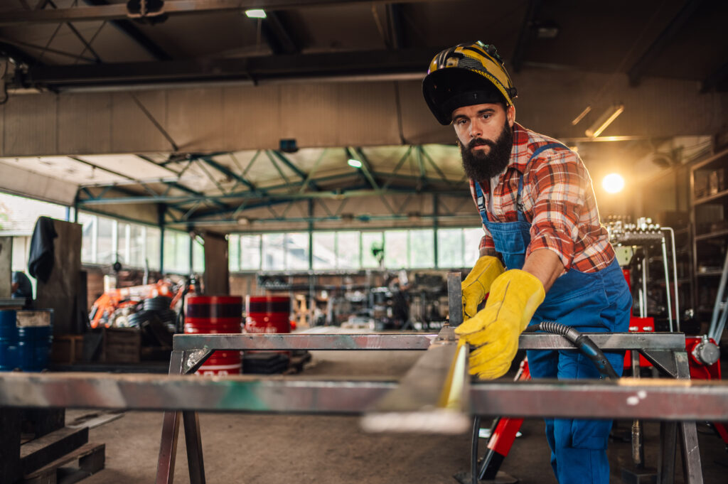 Young bearded man taking precise measurements of the stainless steel while using measuring tape. Lifting his welding mask visor up while marking spots for grinding. Wearing safety equipment.